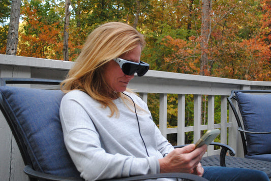 A woman sits outside on a deck, wearing a pair of Eyedaptic EYE5 augmented reality glasses. The lightweight headset is connected to the phone, demonstrating its use for enhanced vision assistance.