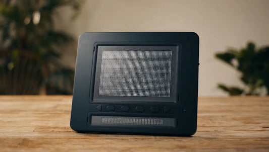 Floridareading Dot Pad, a black and white electronic device with a screen that displays dots and buttons on the bottom. It is sitting on a wooden table.