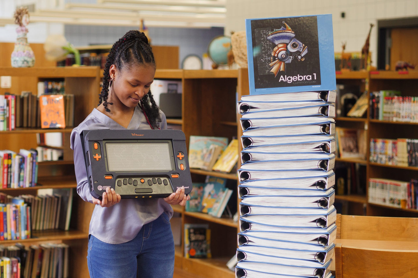 Student holding a Monarch braille display in front of Algebra textbooks in a library.