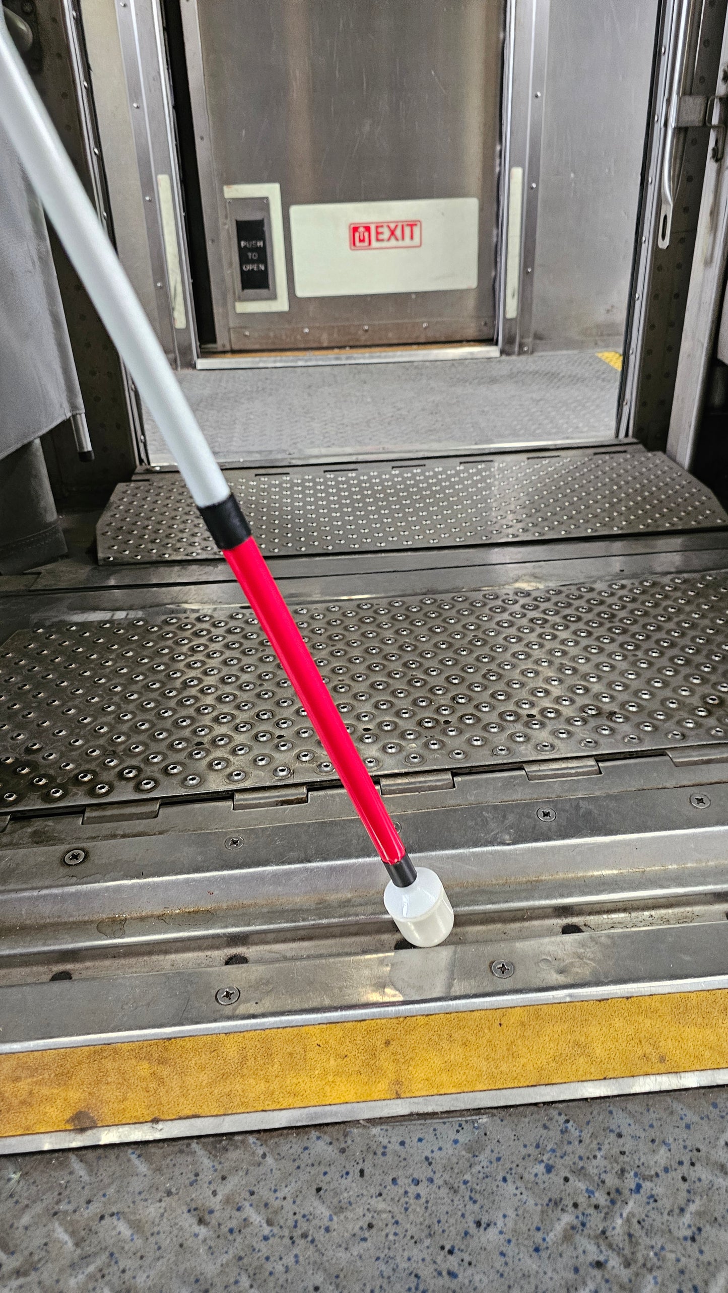 A white cane with a red tip is being used by someone to navigate a train platform. The cane is resting on a textured metal surface and is pointing towards a door with an exit sign visible above it.