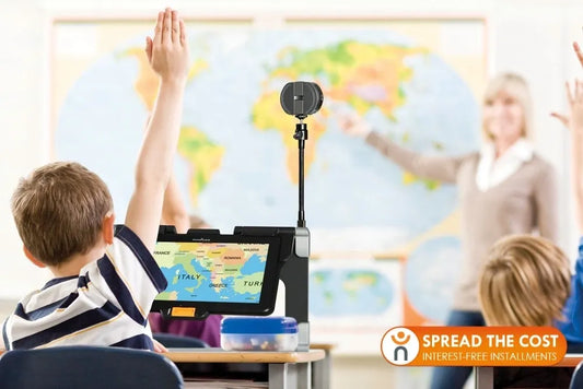 A child in a classroom sits in front of a Prodigi Connect 12 V2.1 tablet with his hand raised. The tablet shows a map of Europe. The teacher stands at the front of the classroom pointing to the map.