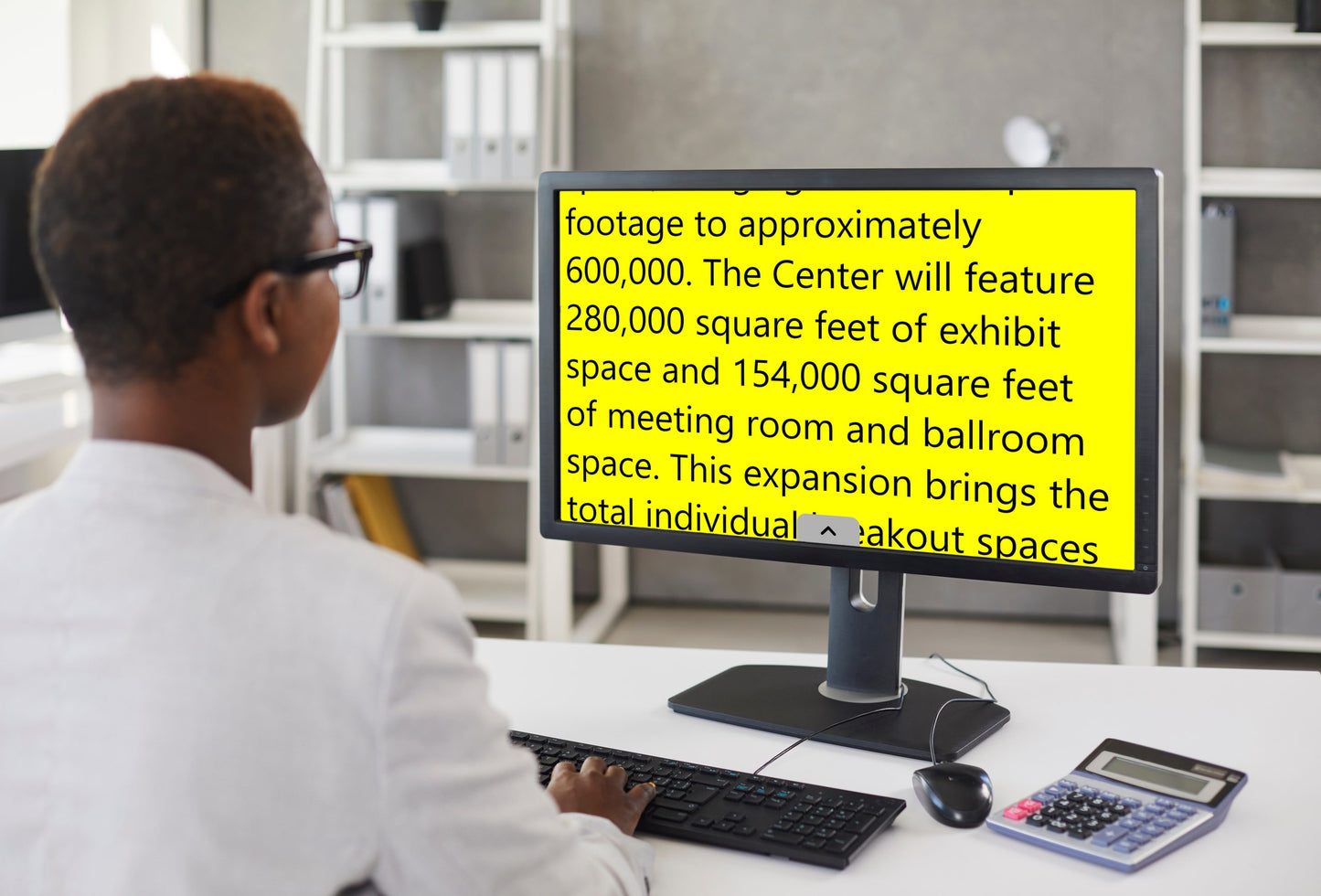 A woman in glasses works at a desk, using Prodigi Software, with a computer screen showing a yellow sign.