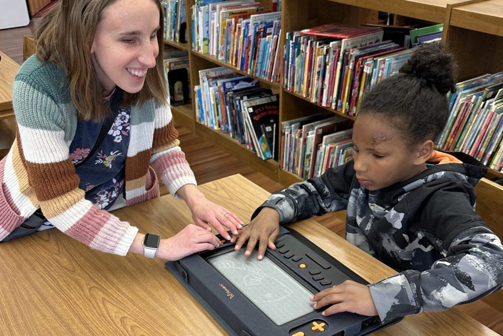 Teacher guiding a child in using the Monarch Braille Display in a library, making braille reading more engaging for young learners.