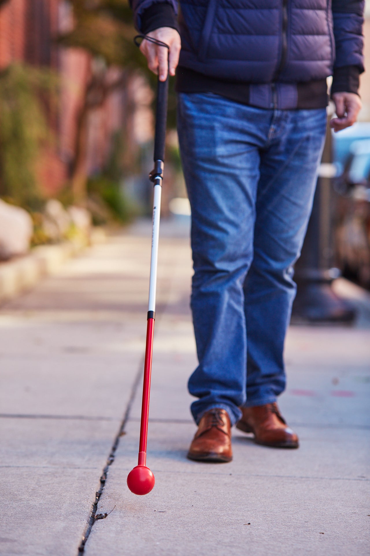 A person with a visual impairment is walking down a sidewalk while holding a white cane with a red tip.