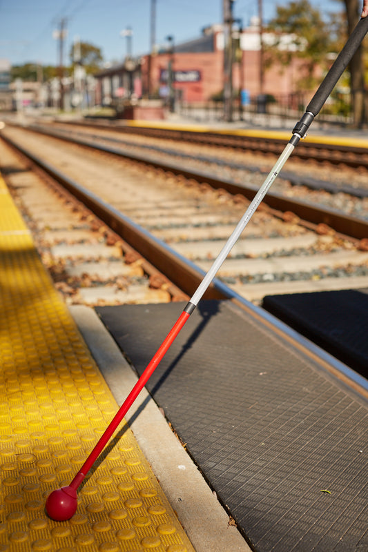 White cane with red tip resting on yellow tactile paving near a train track.