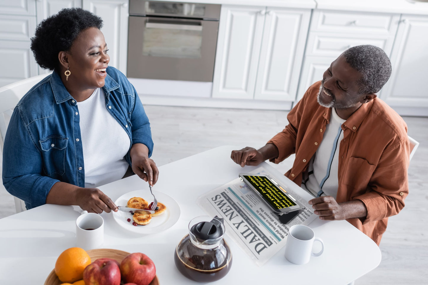 Smiling couple having breakfast, with the man using a digital magnifier to read a newspaper.