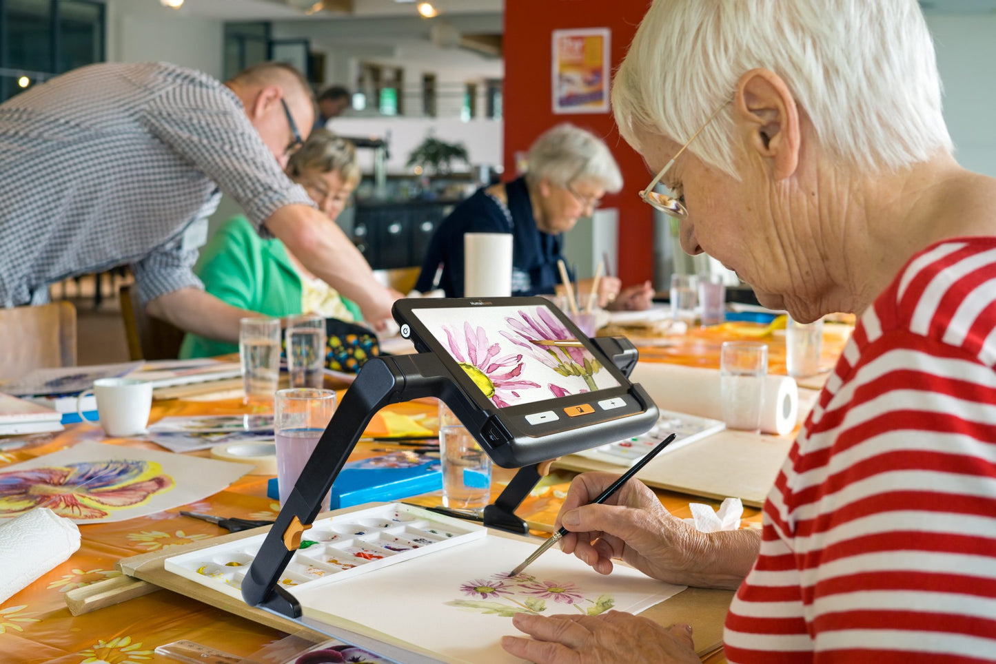 Elderly woman painting flowers while using a digital magnifier in an art class with other seniors.