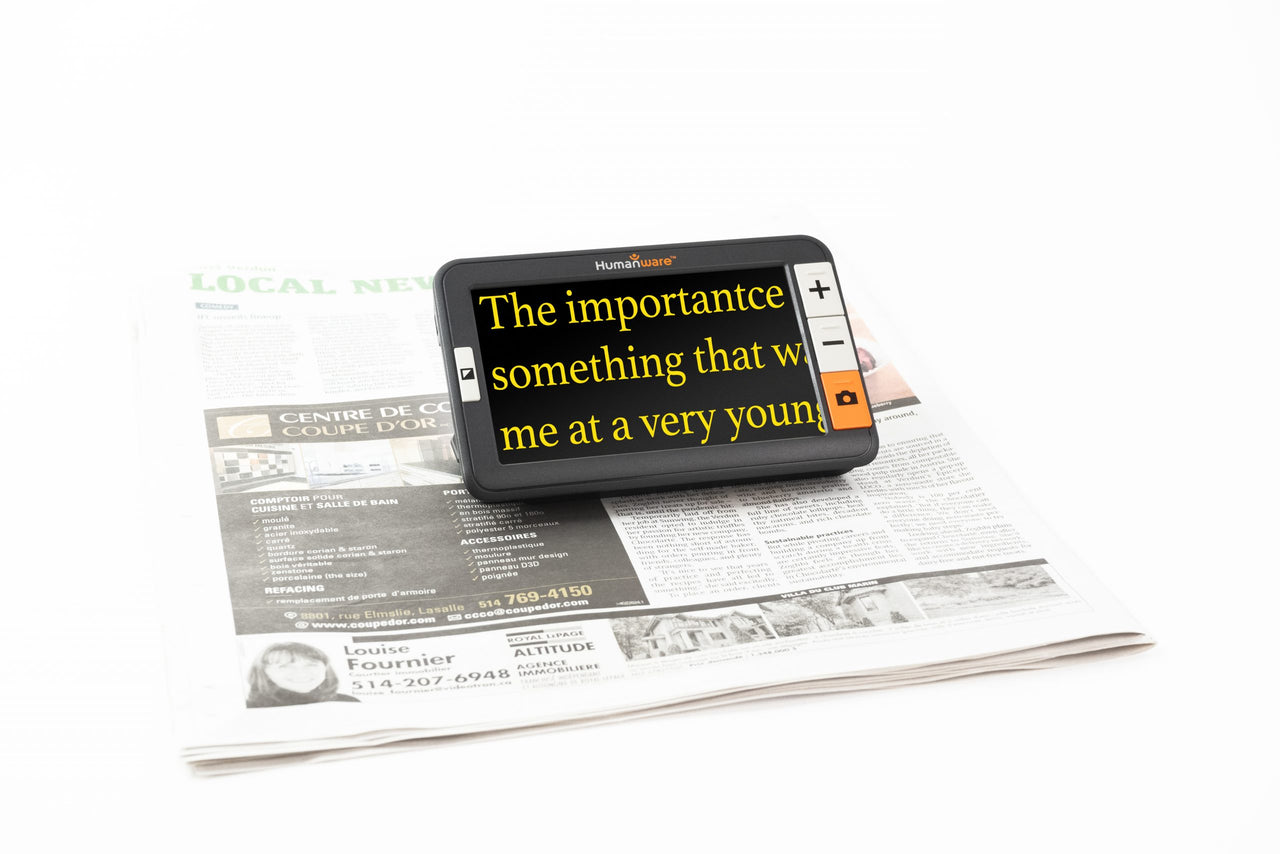 A handheld electronic magnifier with a magnified display showing text from a newspaper. The device has a Humanware logo and controls for zooming and taking photos.